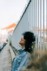 Вид сбоку жизнерадостной афроамериканки в джинсовой куртке, стоящей на тротуаре, опирающейся на забор и улыбающейся — стоковое фото