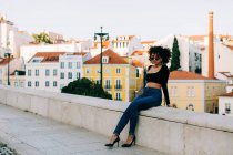 Young trendy African American woman in jeans and crop top sitting on stone parapet and looking over sunglasses — Stock Photo