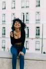 Jeune Afro-Américaine à la mode en jeans et sur le toit d'une récolte assise sur un parapet de pierre et regardant la caméra — Photo de stock