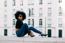 Молодая трепетная афроамериканка в джинсах и кроссовках сидит на каменном парапете и смотрит на солнцезащитные очки — стоковое фото