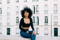 Young trendy African American woman in jeans and crop top sitting on stone parapet and looking at camera — Stock Photo