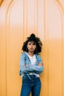 Молодая афроамериканка в джинсах и джинсовой куртке опирается на желтую дверь и смотрит в камеру — стоковое фото