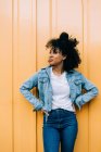 Jeune femme afro-américaine en jeans et veste en denim appuyé sur la porte jaune et détournant les yeux — Photo de stock