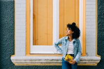 Trendige ethnische Frau in Jeans und Jeansjacke mit modischer Handtasche auf der Fensterbank im Freien — Stockfoto