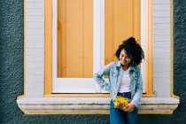 Trendy ethnic woman in jeans and denim jacket holding fashion handbag and leaning on bright windowsill outdoors — Stock Photo