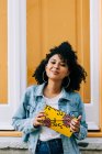 Young African American woman in jeans and denim jacket leaning on yellow door, holding clutch and looking at camera — Stock Photo