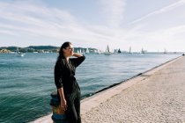 Vista lateral de la joven atractiva mujer en traje negro de pie junto al mar y apoyado en el poste en Lisboa - foto de stock