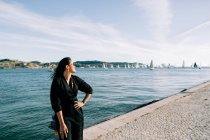Бічний вид на молоду привабливу жінку в чорному одязі, що стоїть на березі моря і спирається на полюс у Лісабоні. — стокове фото