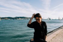 Vue latérale d'une jeune femme séduisante en tenue noire debout au bord de la mer et penchée sur un poteau à Lisbonne — Photo de stock