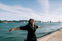 Vue latérale d'une jeune femme séduisante en tenue noire debout au bord de la mer et penchée sur un poteau à Lisbonne — Photo de stock