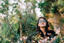 Beautiful brunette woman in glasses standing among blooming bushes in park and looking at camera in Lisbon — стокове фото