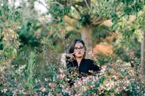 Hermosa mujer morena en gafas de pie entre arbustos florecientes en el parque y mirando a la cámara en Lisboa - foto de stock