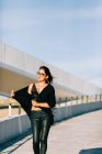 Beautiful stylish woman in black outfit standing with raised hand by cable-stayed bridge in Lisbon on sunny day — Stock Photo