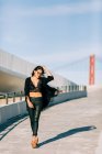 Schöne stilvolle Frau im schwarzen Outfit steht mit erhobener Hand an der Schrägseilbrücke in Lissabon an einem sonnigen Tag — Stockfoto