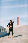 Schöne stilvolle Frau im schwarzen Outfit steht mit erhobener Hand an der Schrägseilbrücke in Lissabon an einem sonnigen Tag — Stockfoto