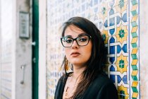 Attraktive junge Frau in Brille und schwarzer Bluse steht an der bunten Vintage-Wand und blickt in Lissabon in die Kamera — Stockfoto