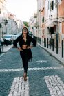 Jeune femme à la mode en tenue noire debout sur le passage supérieur avec les mains sur les hanches à Lisbonne et souriant — Photo de stock
