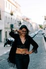 Mujer joven de moda en traje negro de pie en el paso de peatones con las manos en las caderas en Lisboa y sonriendo - foto de stock