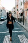 Giovane donna alla moda in abito nero in piedi sul marciapiede con le mani sui fianchi a Lisbona e sorridente — Foto stock