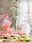Pessoa derramando limonada melancia fria em vidro de frasco na mesa de cozinha rústica ao lado de um monte de flores de gypsophila — Fotografia de Stock