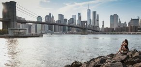 Женщина сидит на камнях в Бруклине и смотрит на мост и Манхэттен в солнечный день — стоковое фото