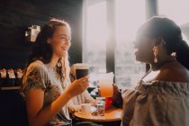 Вид збоку веселі багаторасові молоді випадкові жінки сміються і п'ють каву, сидячи біля вікна в кафе на заході сонця — стокове фото