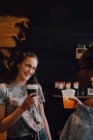 Vista lateral de alegres mujeres casuales jóvenes multirraciales riendo y bebiendo café mientras se sienta junto a la ventana en la cafetería al atardecer - foto de stock
