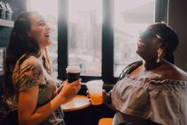 Side view of cheerful multiracial young casual women laughing and drinking coffee while sitting by window at cafe on sunset — Stock Photo