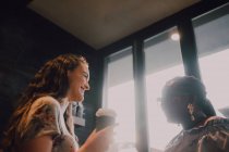 D'en bas des jeunes femmes occasionnelles multiraciales joyeuses riant et buvant du café tout en étant assis par la fenêtre au café au coucher du soleil — Photo de stock
