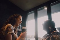 From below of cheerful multiracial young casual women laughing and drinking coffee while sitting by window at cafe on sunset — Stock Photo