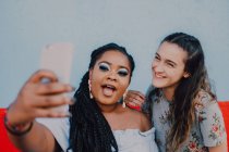 Multirazziale giovani donne casual ridere e scattare selfie con smartphone su sfondo chiaro — Foto stock