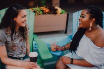 Cheerful multiracial young casual women talking and drinking coffee while sitting by vibrant table at hipster cafe — Stock Photo