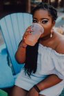 Alegre afro-americano jovem mulher bebendo bebida fria enquanto sentado no café hipster, olhando para longe — Fotografia de Stock