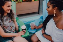 Cheerful multiracial young casual women talking and drinking coffee while sitting by vibrant table at hipster cafe — Stock Photo