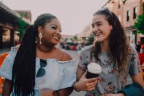 Fröhliche trendige multirassische junge Frauen, die sich unterhalten und Kaffee trinken, während sie bei Sonnenuntergang auf der Straße spazieren gehen — Stockfoto