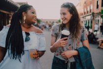 Fröhliche trendige multirassische junge Frauen, die sich unterhalten und Kaffee trinken, während sie bei Sonnenuntergang auf der Straße spazieren gehen — Stockfoto