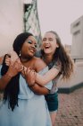 Multiracial young casual women laughing and hugging while standing on street on sunset — Stock Photo