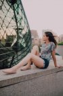 Calma contenido casual joven mujer en pantalones cortos y camiseta disfrutando de la luz del sol mientras está sentado en parapeto de hormigón, mirando hacia otro lado - foto de stock