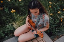 Souriant tendance casual jeune femme en t-shirt jouer ukulele tout en étant assis sur le trottoir à côté du lit de fleurs — Photo de stock