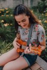 Sorrindo na moda casual jovem em t-shirt jogando ukulele enquanto sentado no pavimento ao lado de canteiro de flores — Fotografia de Stock