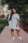 Schwarze junge Frau mit langen Zöpfen im schulterfreien Kleid auf Betonpfad im blühenden Garten und blickt in die Kamera — Stockfoto