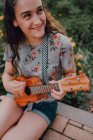 Улыбающаяся модная девушка в футболке, играющая на укулеле, сидя на тротуаре рядом с клумбой — стоковое фото
