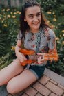 Sorrindo na moda casual jovem em t-shirt jogando ukulele enquanto sentado no pavimento ao lado de canteiro de flores — Fotografia de Stock
