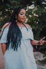 Привлекательная черная молодая женщина с ярким макияжем в платье без плеча слушает музыку на смартфоне с наушниками — стоковое фото