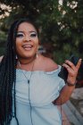 Attraente giovane donna nera con trucco luminoso in abito da spalla ascoltando musica su smartphone con auricolari — Foto stock