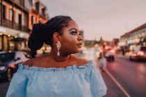 Jolie jeune femme noire avec un maquillage lumineux en robe hors épaule debout sur la rue au coucher du soleil, regardant ailleurs — Photo de stock