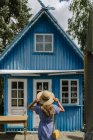 Vista posteriore della donna in cappello di paglia e prendisole in piedi di fronte alla colorata casa estiva blu alla luce del giorno — Foto stock
