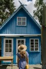Back view of woman in straw hat and sundress standing in front of colourful blue summer house in daylight — Stock Photo