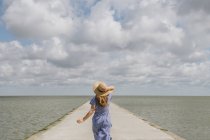 Back view of adult woman in straw hat and sundress running on empty concrete quay on cloudy day — Stock Photo