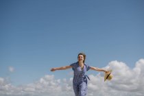 Mujer adulta contenta con el pelo que sopla y en sundress que camina con sombrero de paja en la mano en el fondo nublado del cielo - foto de stock